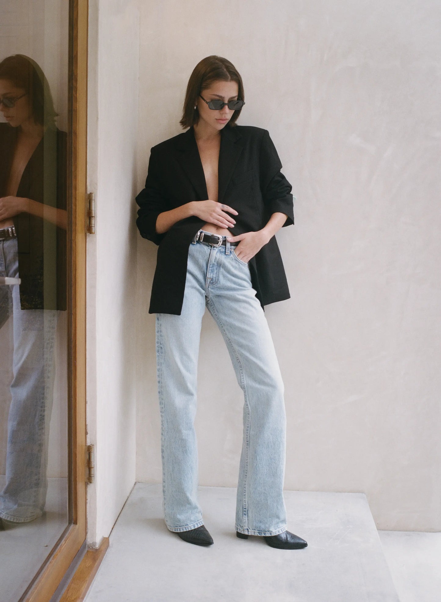 Woman wearing a black blazer and light blue jeans standing in a minimalistic room.