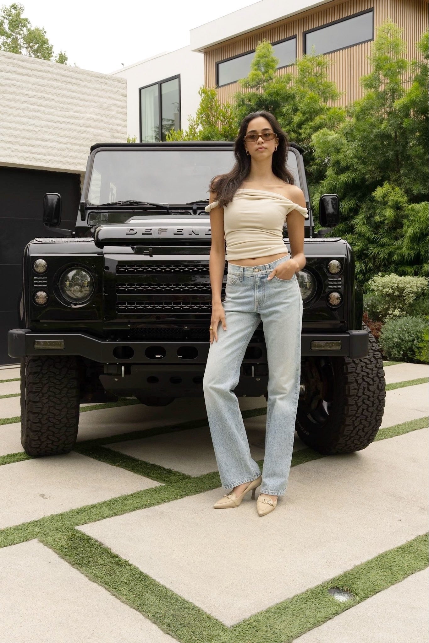 Woman standing in front of a black Land Rover vehicle on a driveway, wearing a white top and lkblue women bootcut denim jeans.
