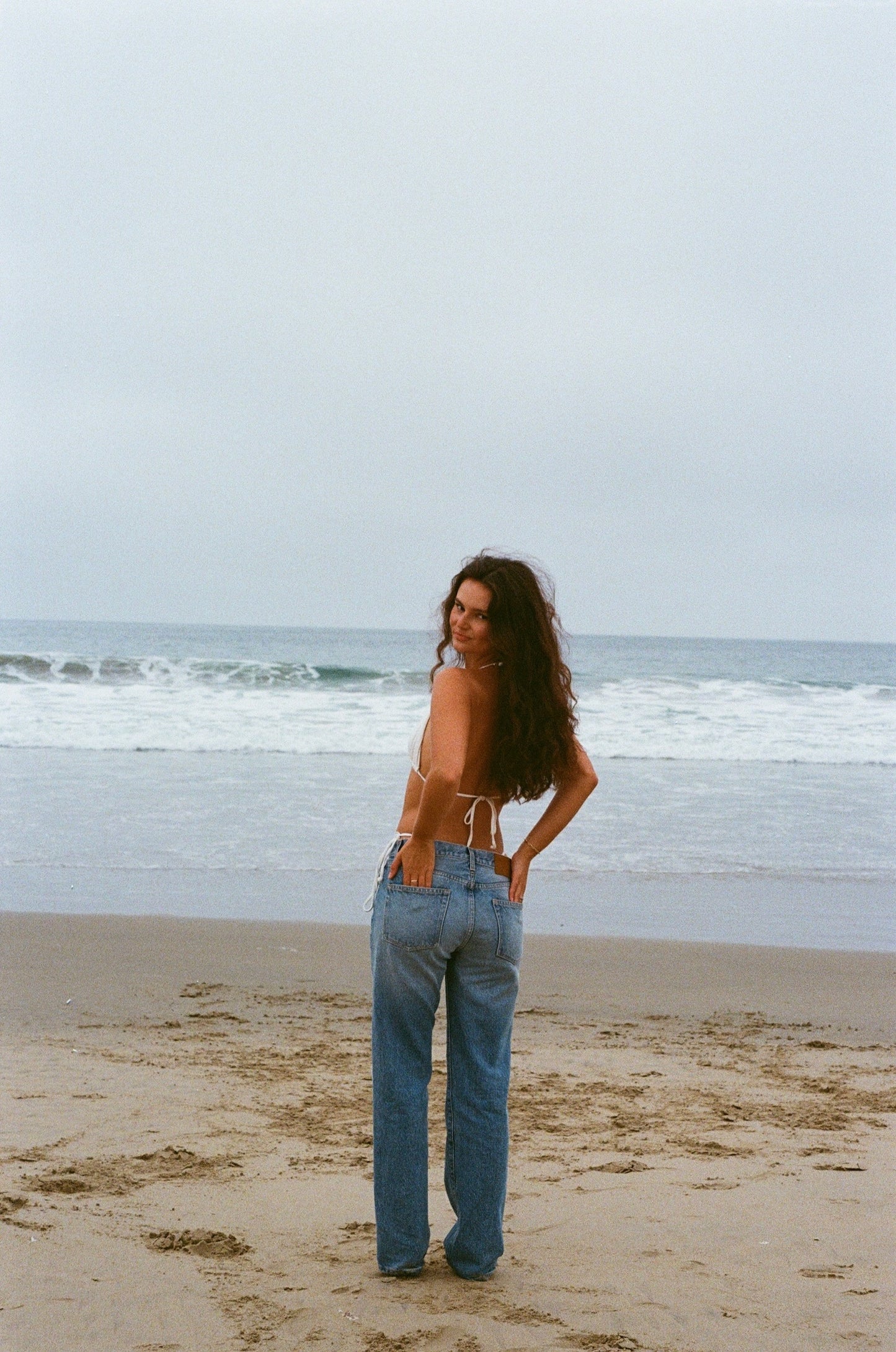 A woman wearing lkblue straight leg jeans of denim standing on a beach with ocean waves in the background