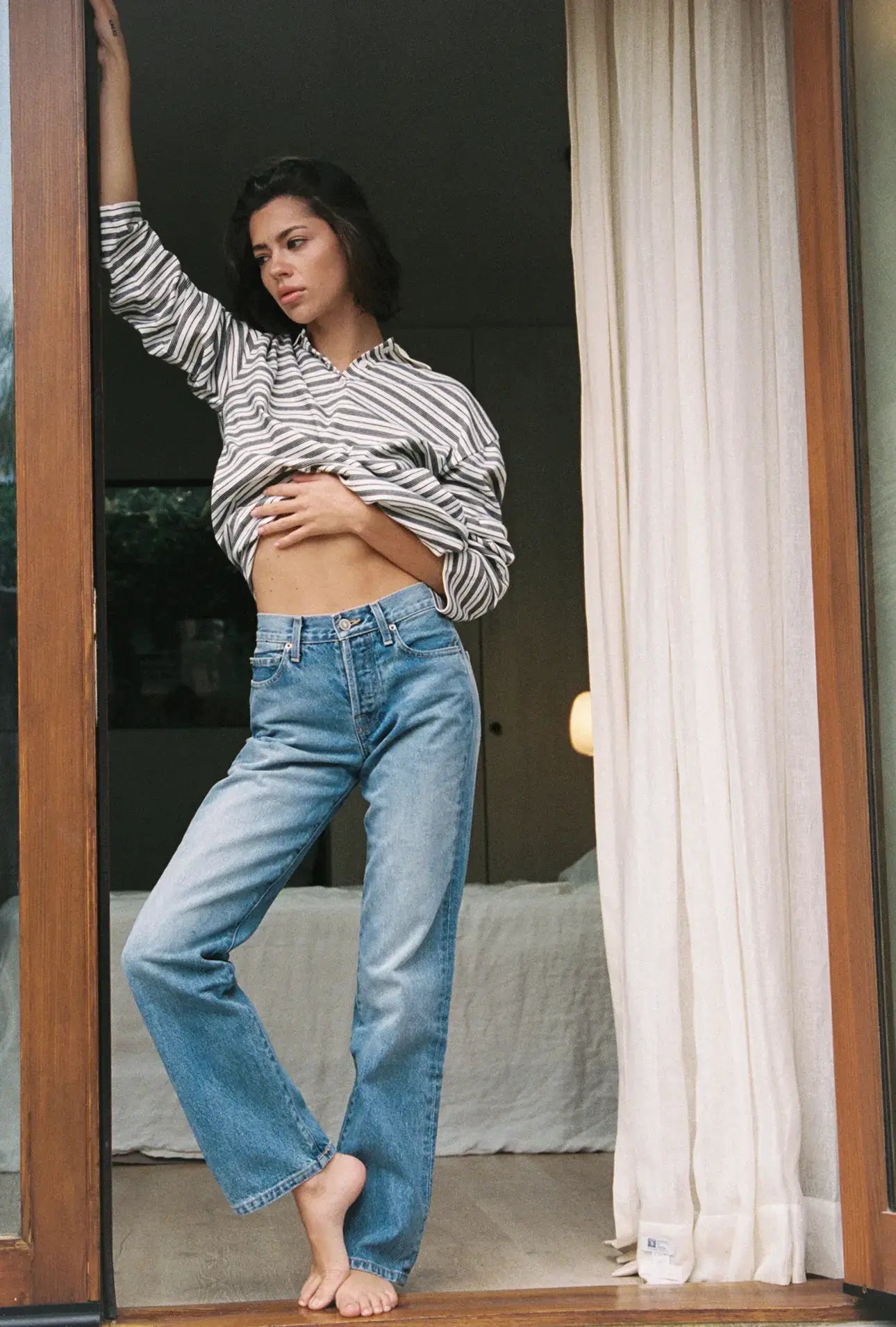 A woman wearing a striped shirt and blue straight leg jeans is standing in front of a glass door with curtains.
