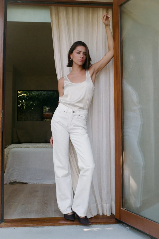 Woman in a white top and white BEL AIR bootcut jean standing in a doorway with curtains and a bed in the background