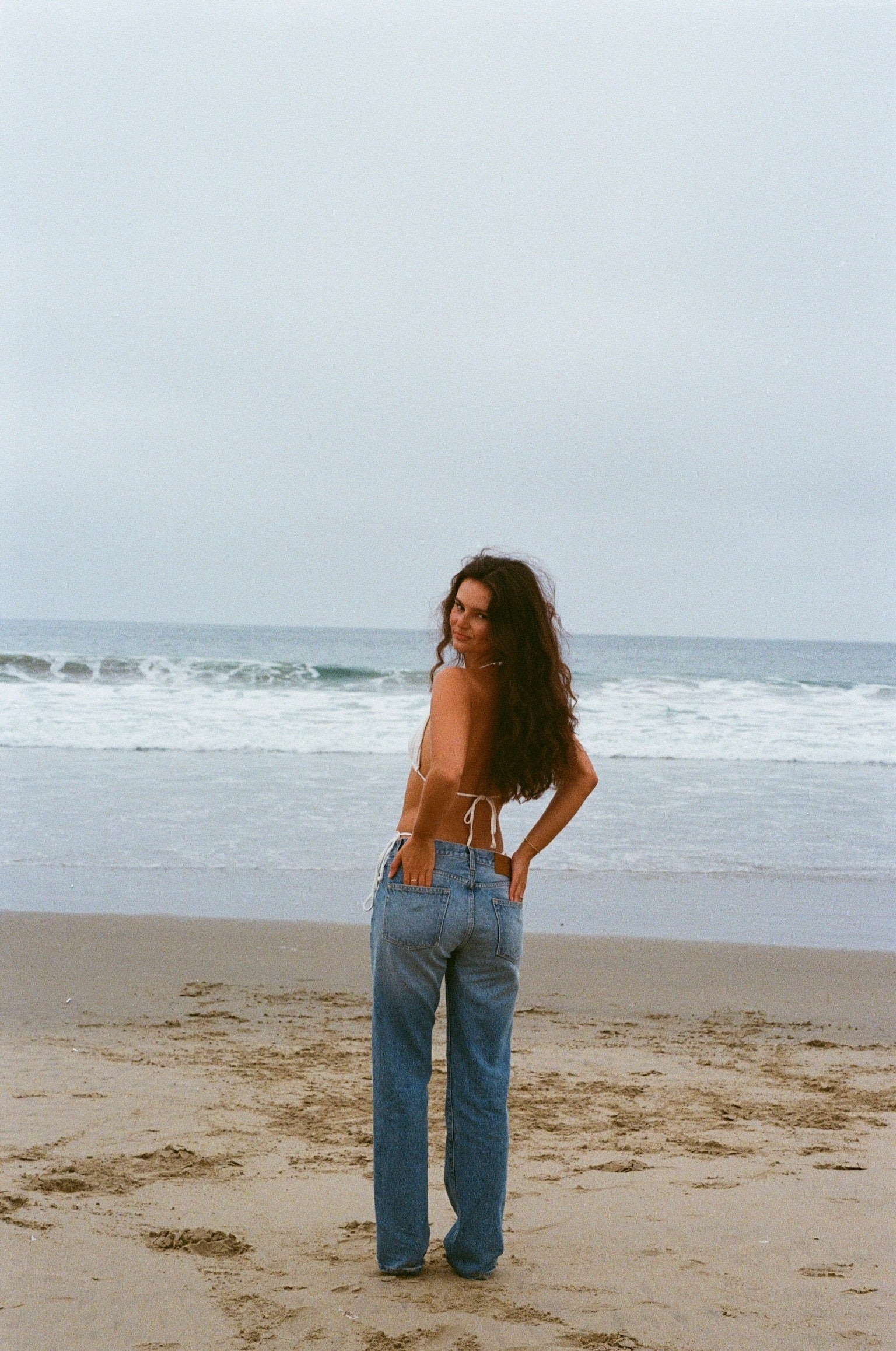 A woman wearing lkblue straight leg jeans of denim standing on a beach with ocean waves in the background