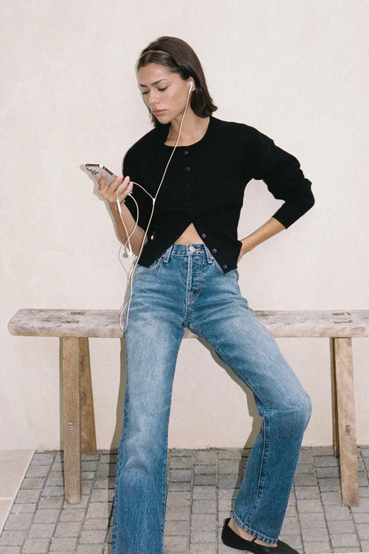 Women in black cropped cardigans and blue denim jeans sit on a wooden bench, listening to music.