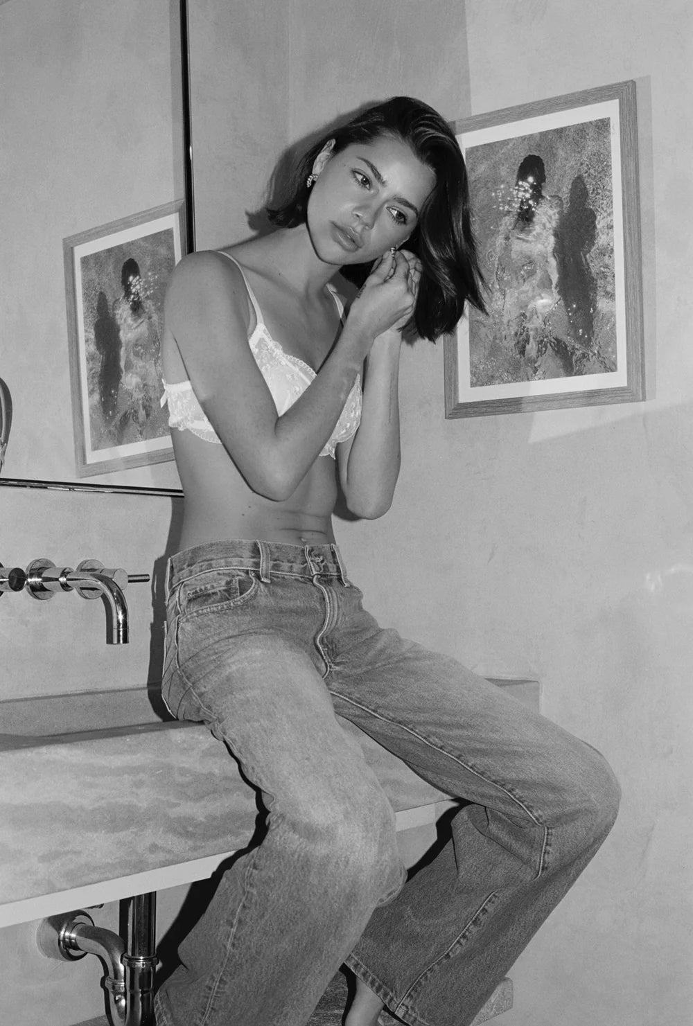 Person sitting on a sink wearing a bra and jeans in a bathroom setting.