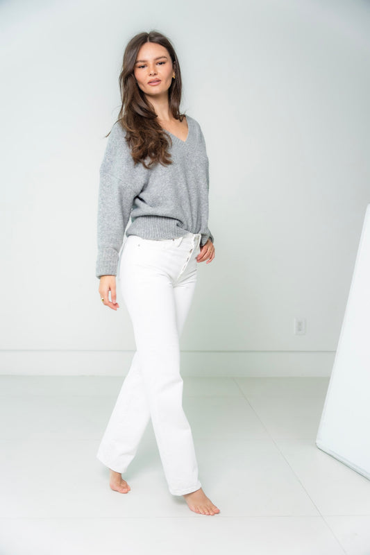 A woman wearing a gray sweater and white straight leg denim  pants against a white background